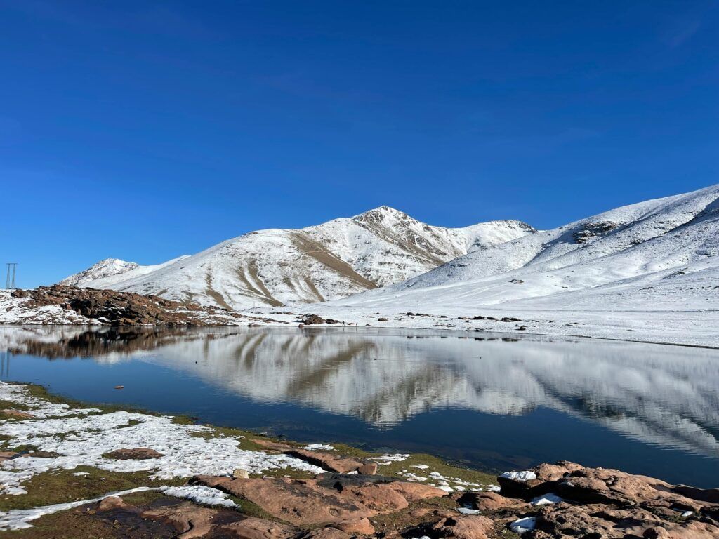 Il momento migliore per visitare il Marocco: una guida locale per pianificare il tuo viaggio Stunning view of snow-capped mountains reflected in a serene alpine lake under a clear blue sky.